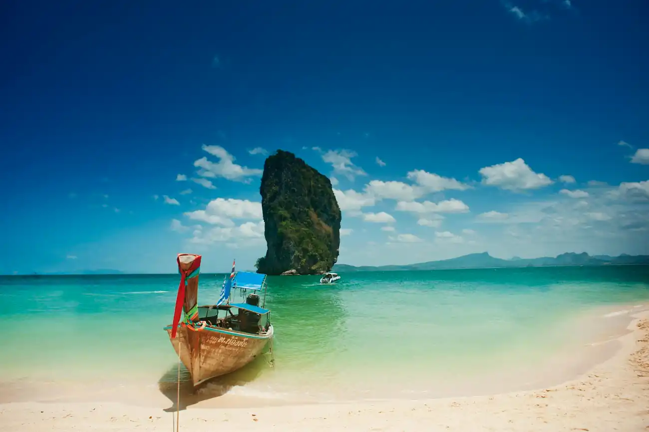 A traditional wooden longtail boat is moored to the sandy beach. Clear blue water extands to the horizon, A blue and white speedboat cruises past a small but high vegetation covered limestone pillar island. In the background is a landscape of cliffs and ridges