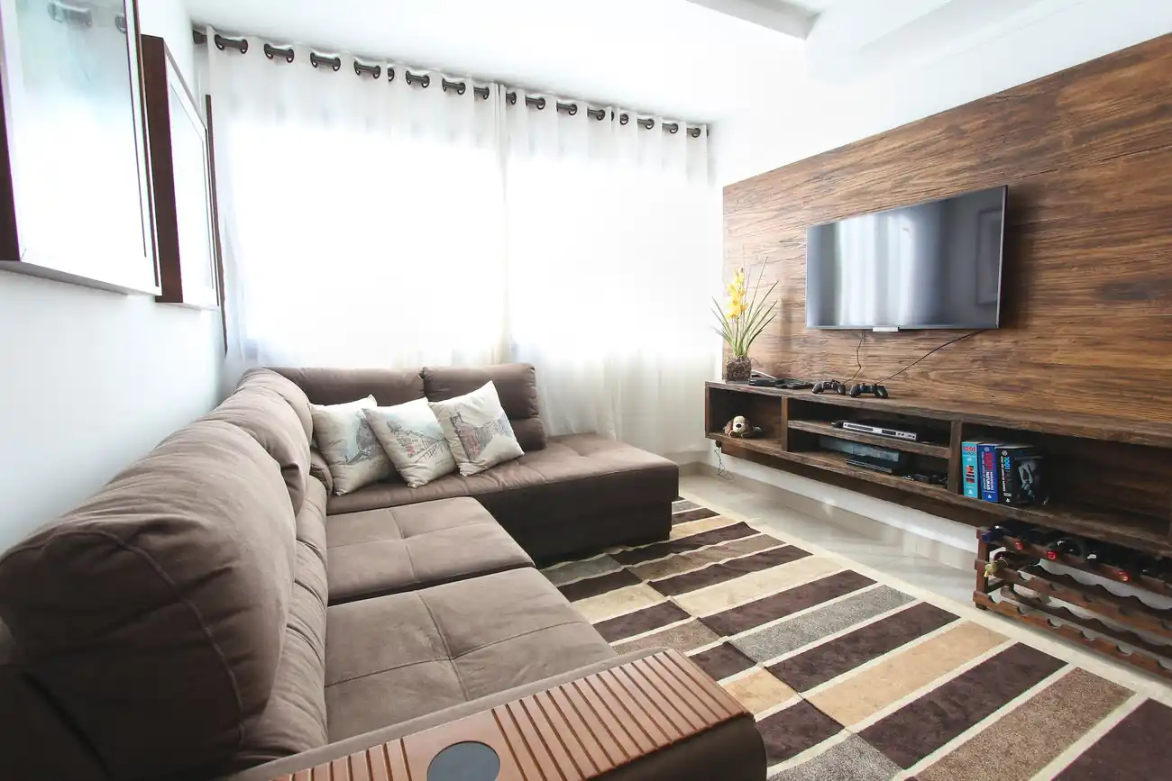 The living room of a condominium. Decor is mostly browns and creams. Featuring a wood paneled wall with a wide screen TV attached. There are game controllers and a wine rack. The far wall is a wide window. White curtains are closed against the sunlight. Opposite the TV, against the wall is an L shaped sofa