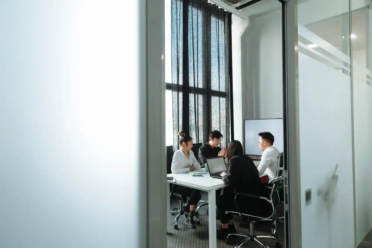 Two women and two men sit around a table, conversing, they are in a corporate meeting room.