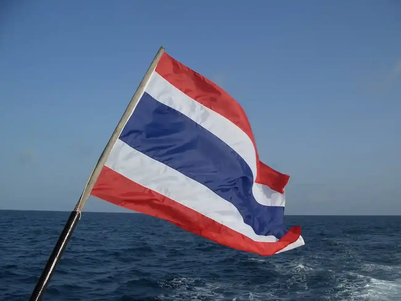 The flag of Thailand unfurled and fluttering in the breeze on a small pole on the stern of a boat. In the background a clear blue sky an open sea to the horizon disturbed only by the wake of the boat we cannot see
