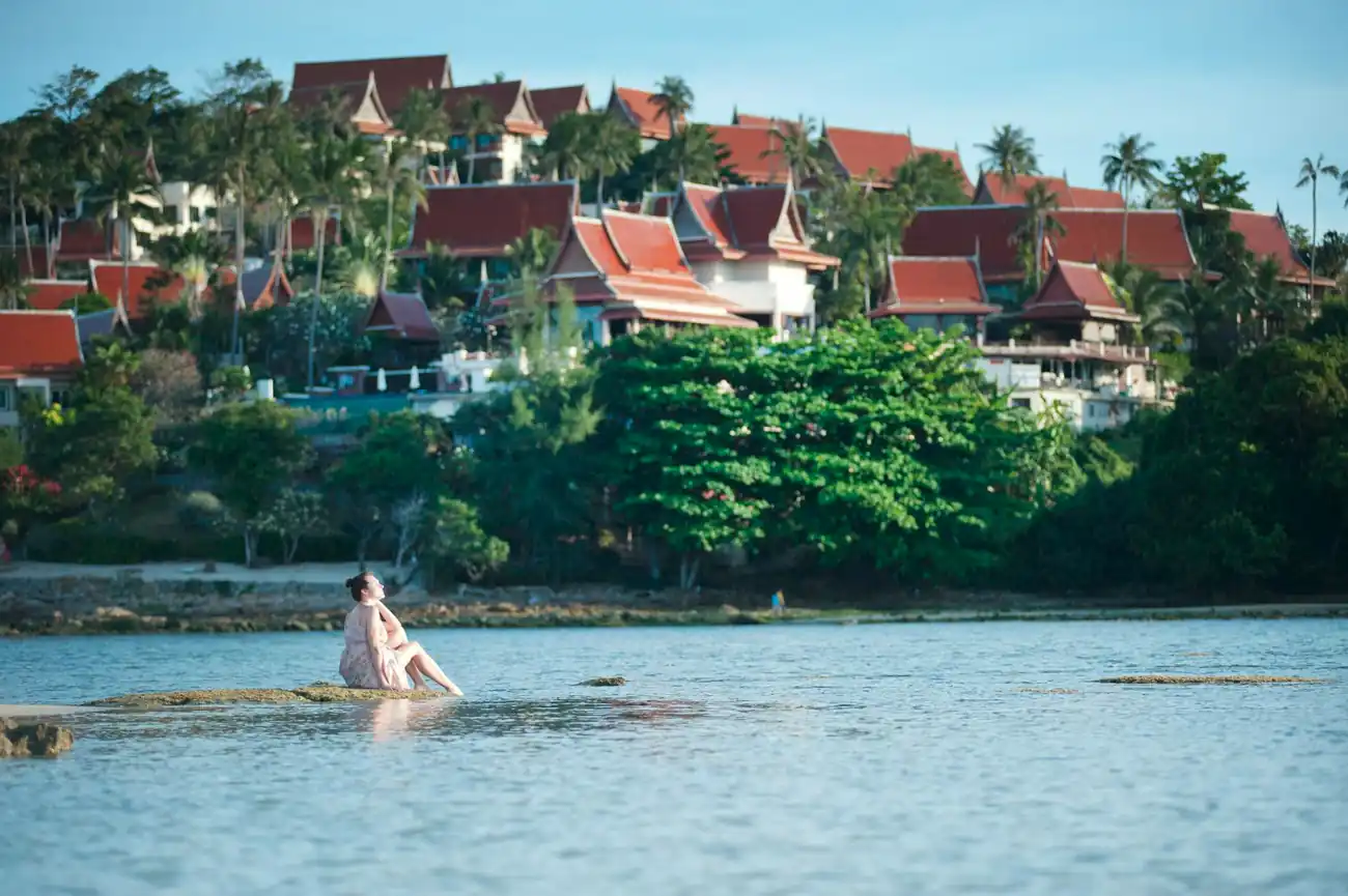 A woman in a dress looks out to sea. She's sitting on a low rock almsot surrounded by shallow water, in the background is a narrow beach and a headland where many Thai styles home have been built. Each iin the same style of white walls and sweeping, curved, red tiled roofs.