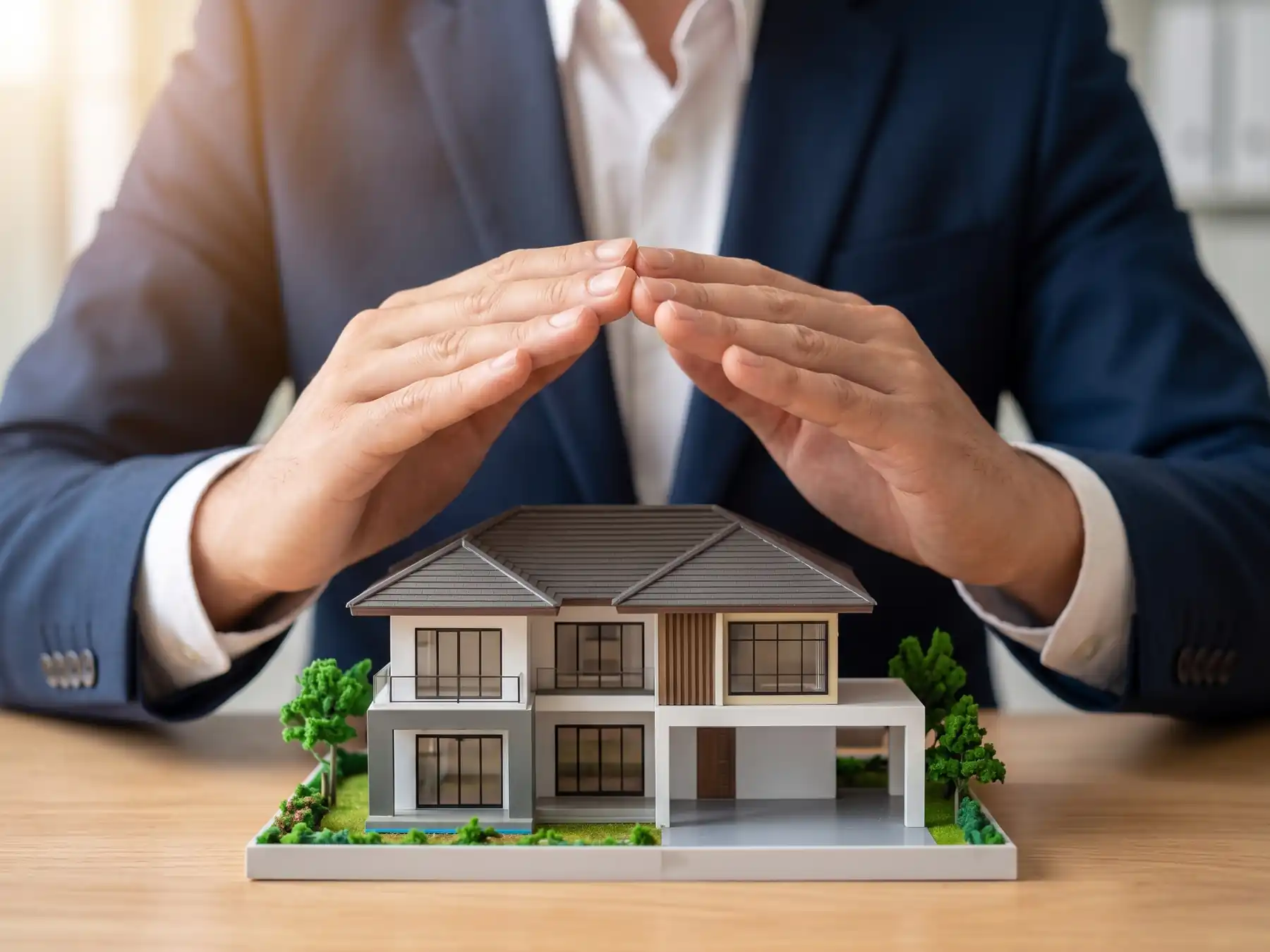 The model of a large residential house is on a desk. Behind we can see the shoulders and chest of a man sitting at the desk wearing a shirt and business jacket. He is cupping his hands over the model symbolising protection of property and assets yu insurance.